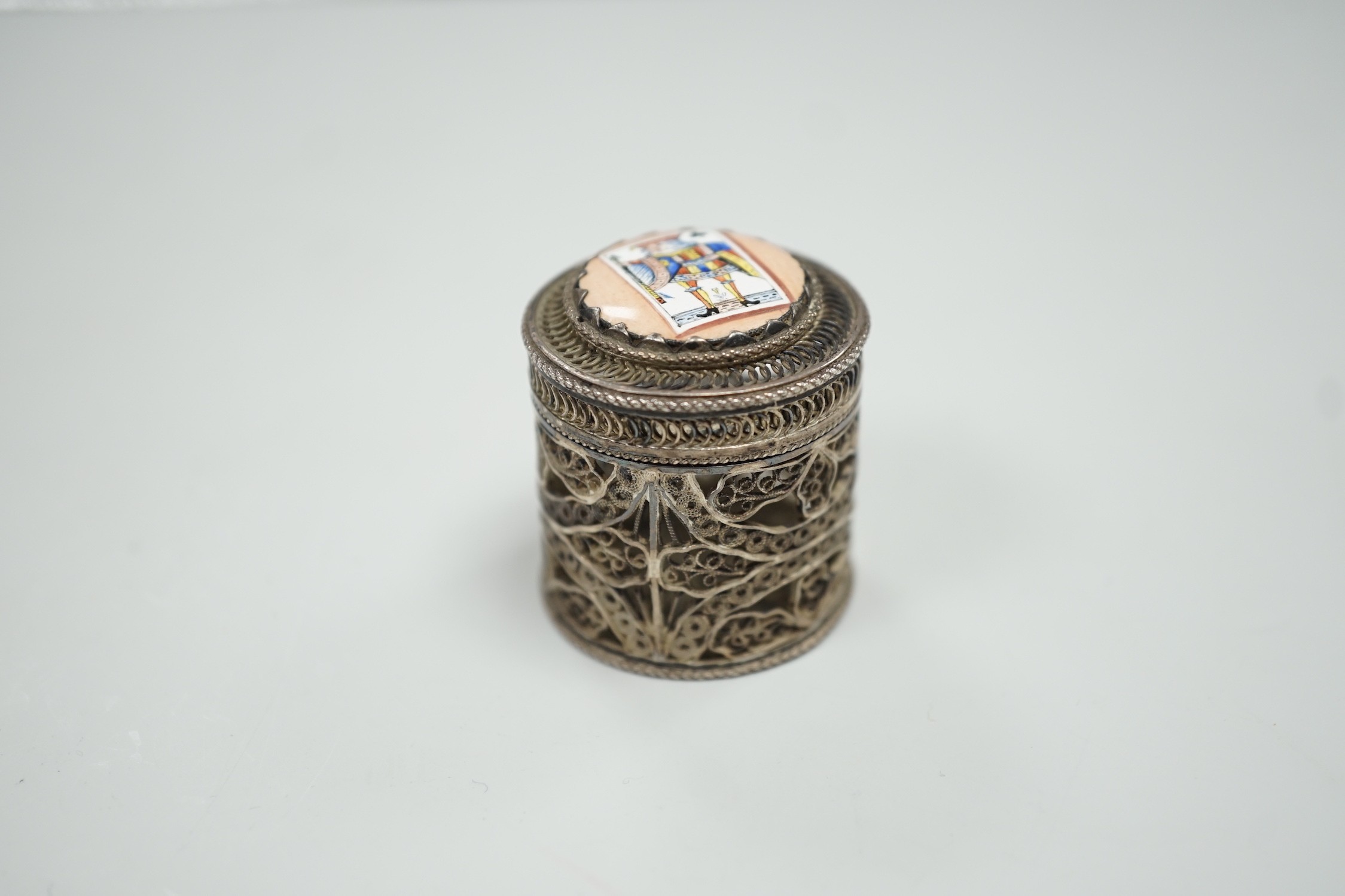 A continental filigree white metal and enamelled cylindrical pill box, the cover enamelled with a picture playing card, height 32mm.